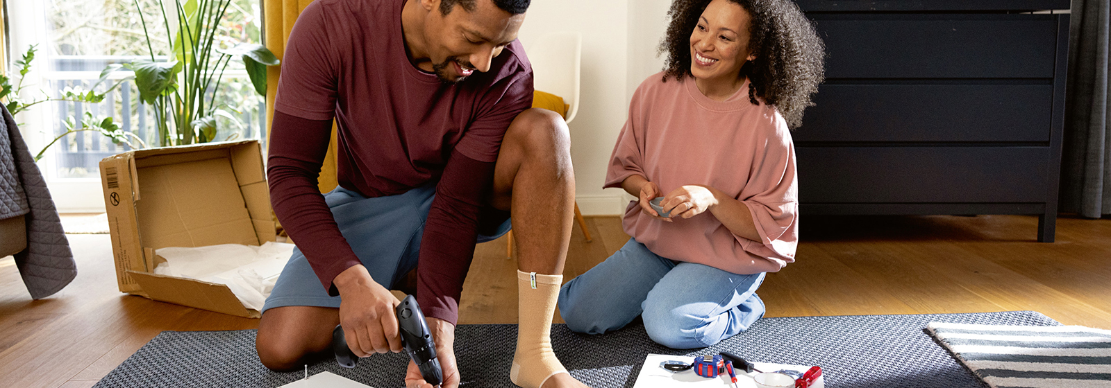 Mann som har på ankelstøtte mens han "gjør-det-selv" skrur skruer på baksiden av et kvadratisk bord mens en kvinne ser på Mann som har på ankelstøtte mens han "gjør-det-selv" skrur skruer på baksiden av et kvadratisk bord mens en kvinne ser på
